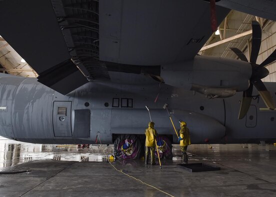 Photo shows Airmen, dressed in yellow protective gear, wetting sponges to clean a C-130J Super Hercules aircraft.