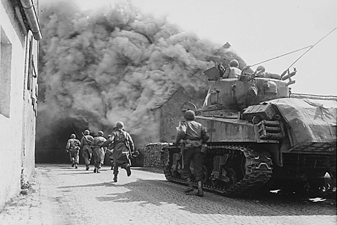 Soldiers run alongside a tank. Smoke billows off a burning home.