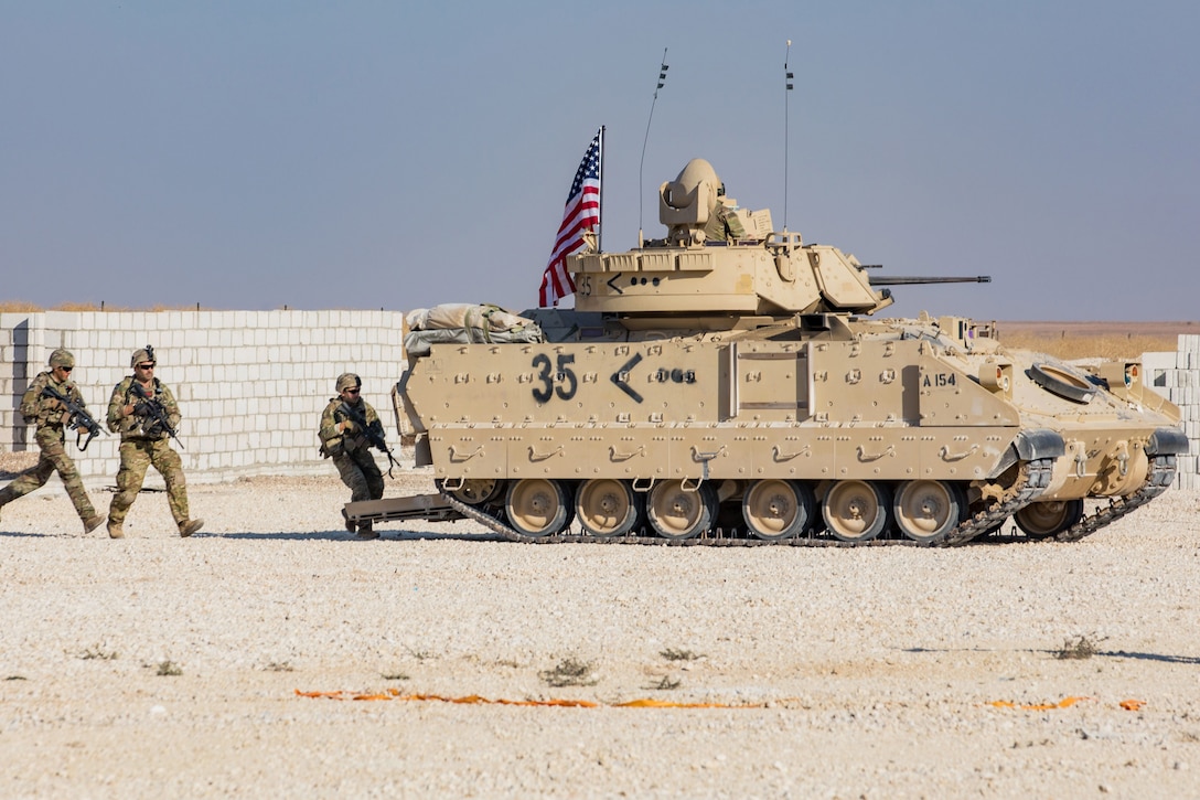 Soldiers run toward a military vehicle on desert terrain.
