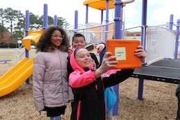 One child holds up mobile tablet to take a selfie, while four friends smile in background