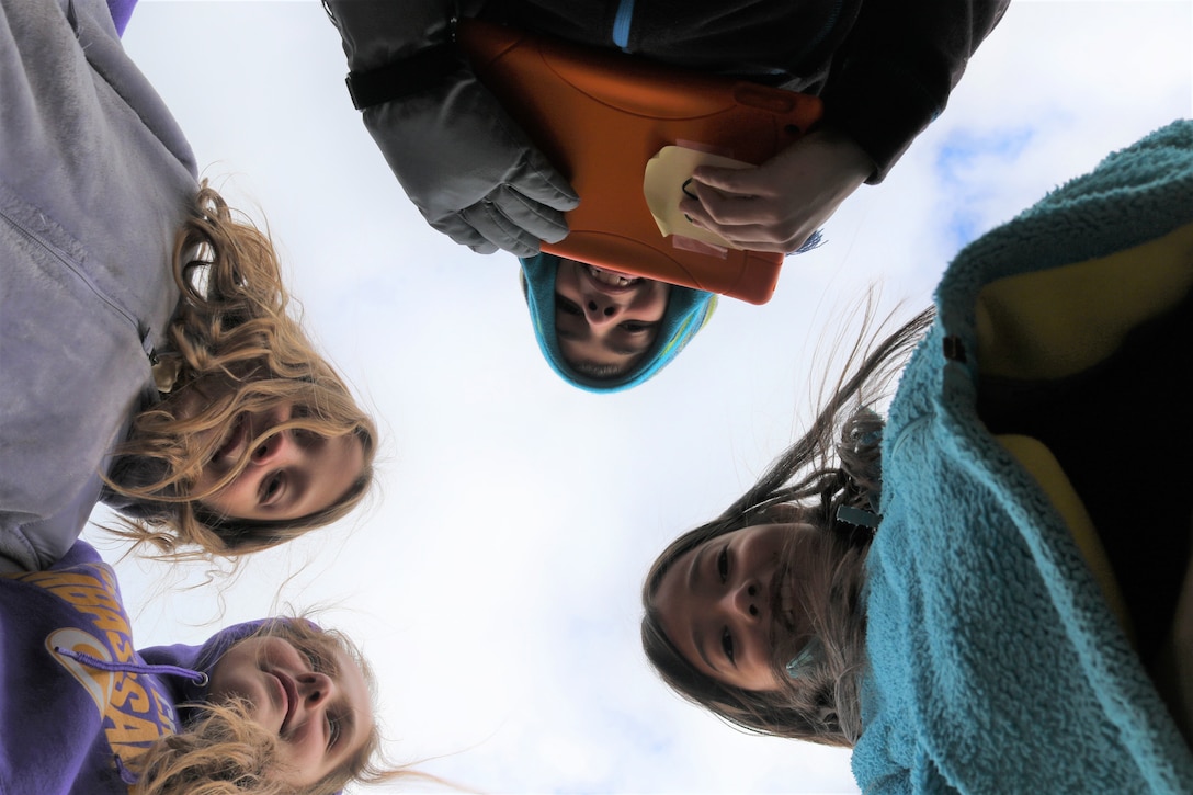 Four young students look down at camera while one holds a mobile tablet