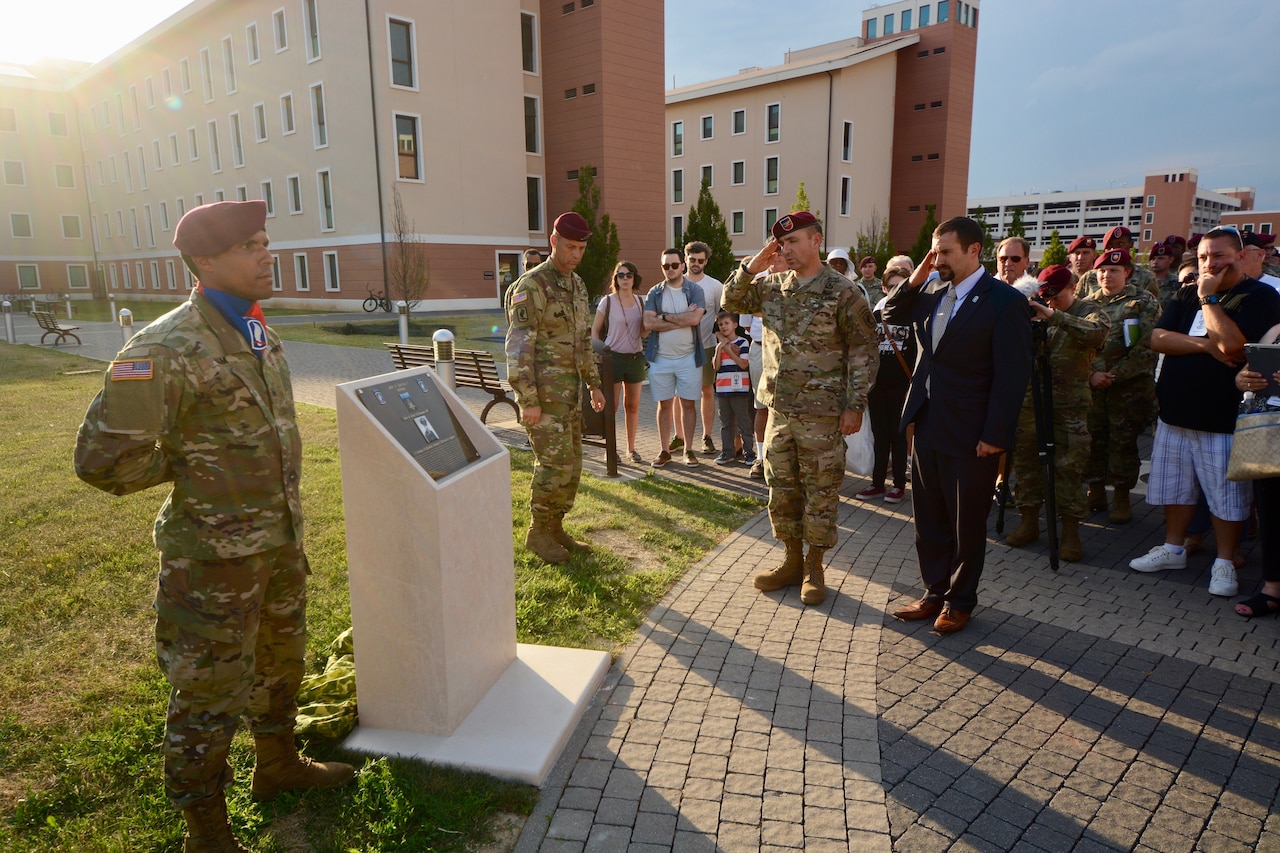 An Army commander and a veteran in plain clothes salute a memorial plaque on a walkway. Other soldiers stand at attention around it. Civilians also look on.