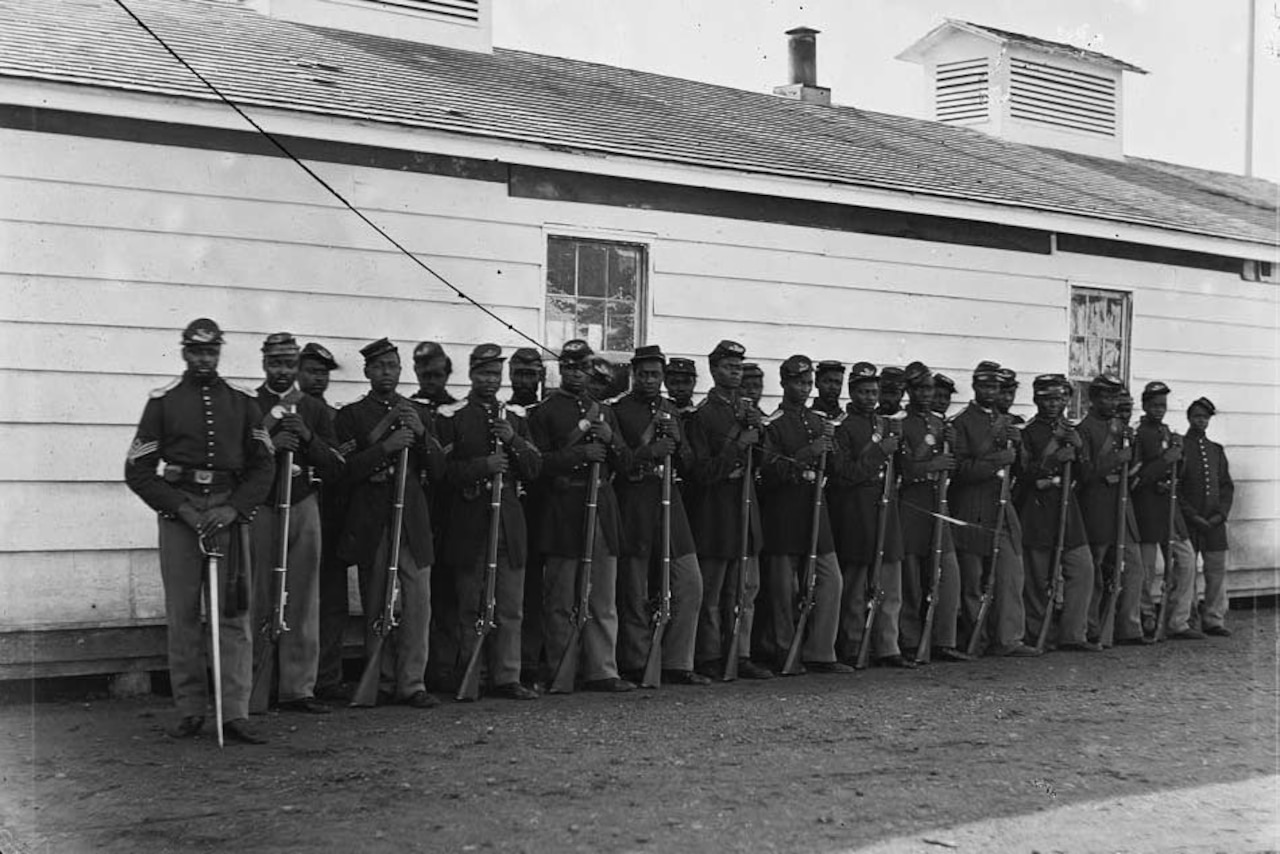 More than two-dozen black Civil War soldiers stand in two lines with their rifles resting on the ground in front of a white building.