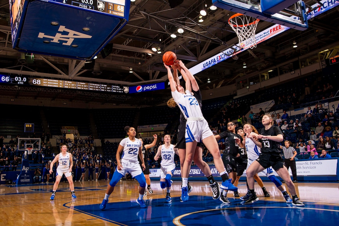 A service member jumps up to grab a basketball will others surround her on a basketball court.