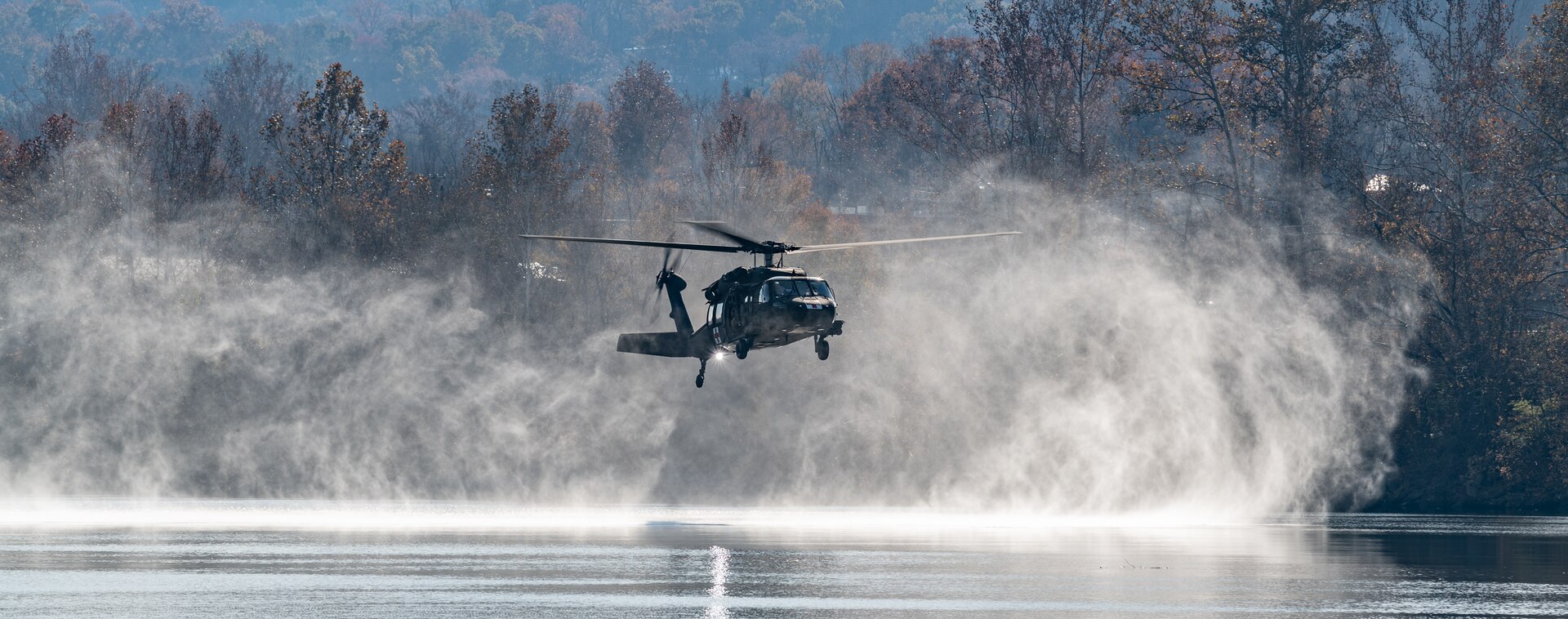 A West Virginia Army National Guard UH-60M Blackhawk helicopter flys low, close to river surface, creating a misty cloud around the helicopter.