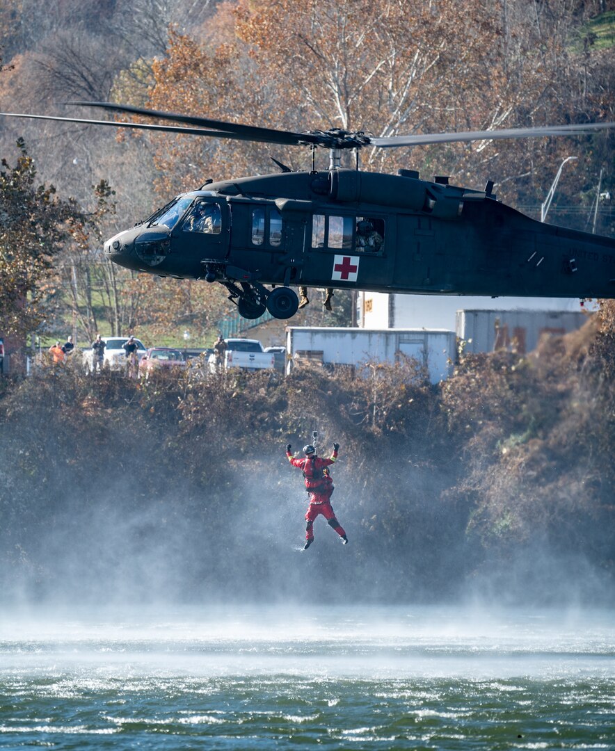A side view of a West Virginia National Guard Blackhawk Helicopter airlifting a participant wearing an orange wet suit out of the Kanawha River.