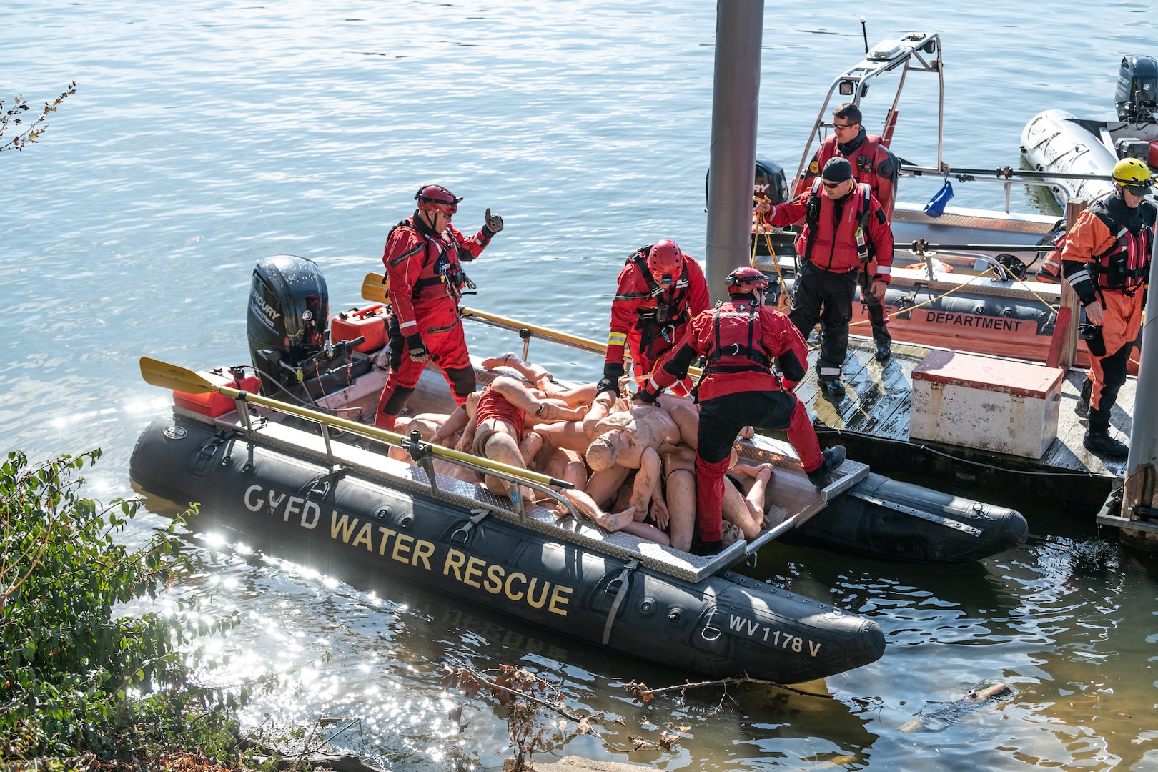 Swift water crew members in red wet suits and gear load mannequins into their boat prior to placement along the banks of the Kanawha River.