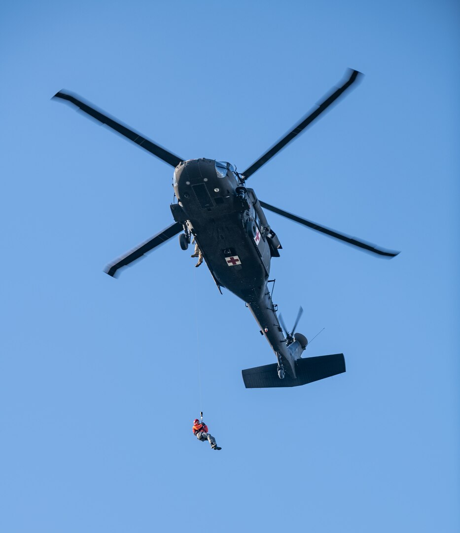 From below, a West Virginia Army National Guard UH-60M Blackhawk helicopter flying while carrying a participant on a hoist line against a bright blue sky.