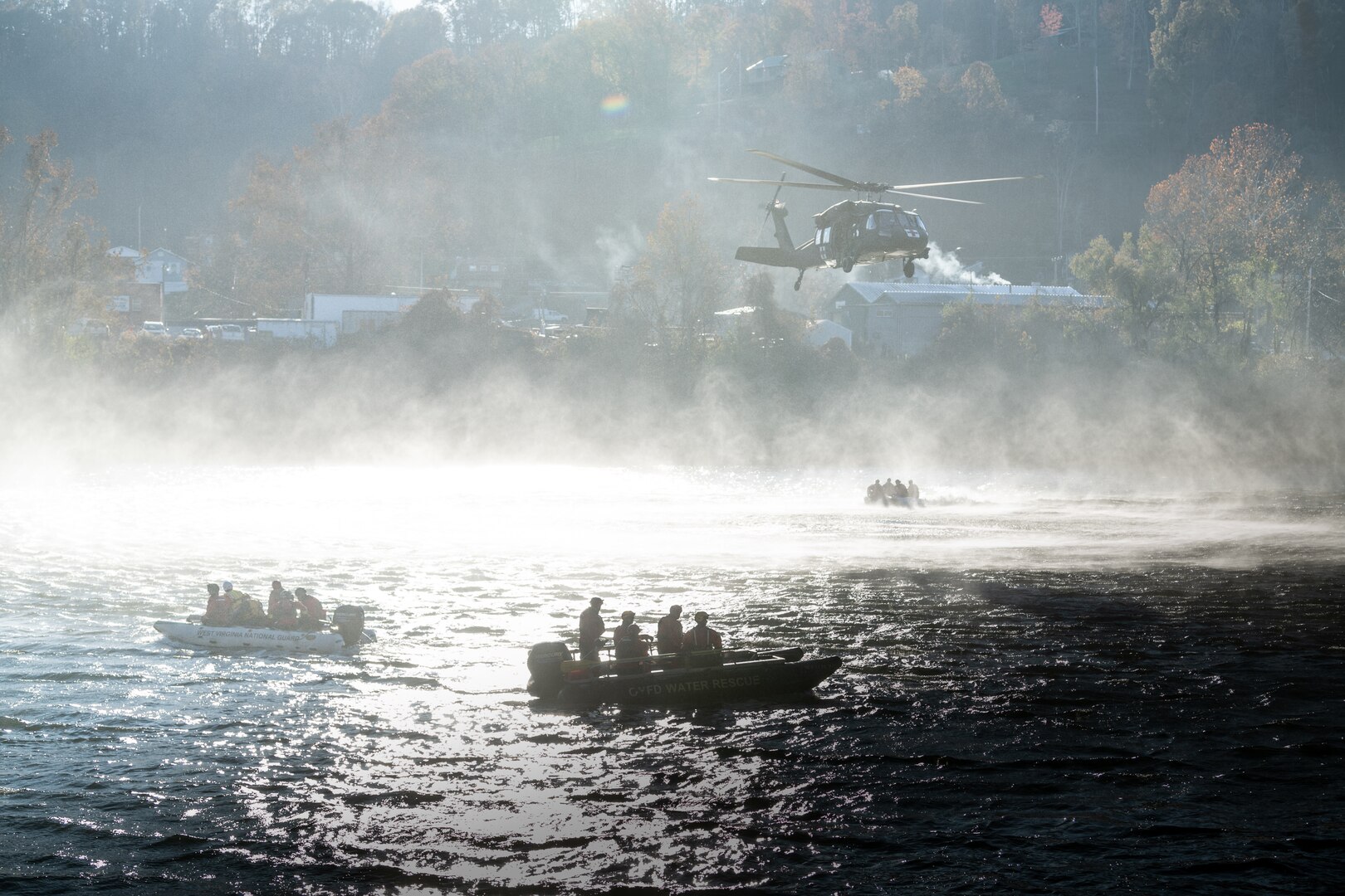 Three swift water boats below a Blackhawk helicopter, in a silhouette with the sun behind and rotor wash creating a misty fog across the river.