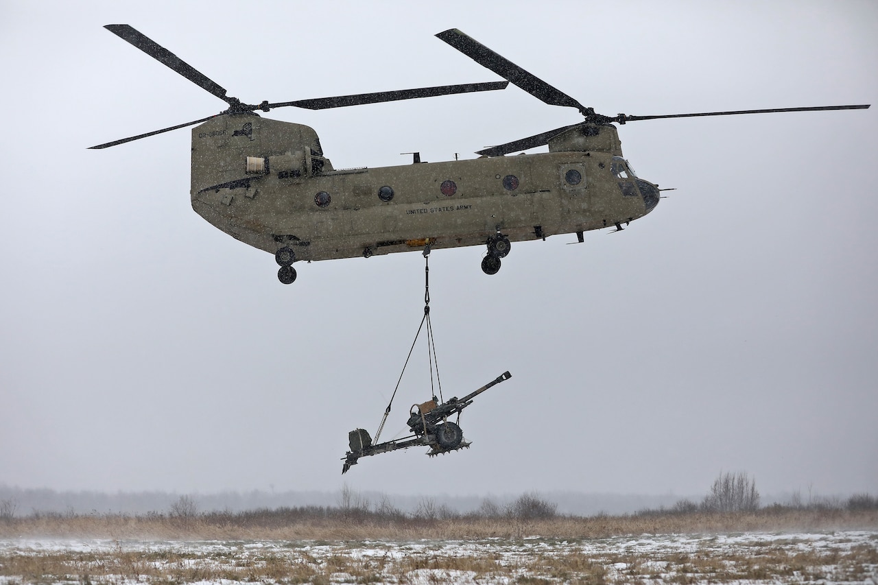 A helicopter hovers with a howitzer hanging by a sling.