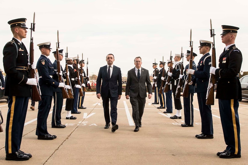 Two men in suits walk between two rows of uniformed service members.