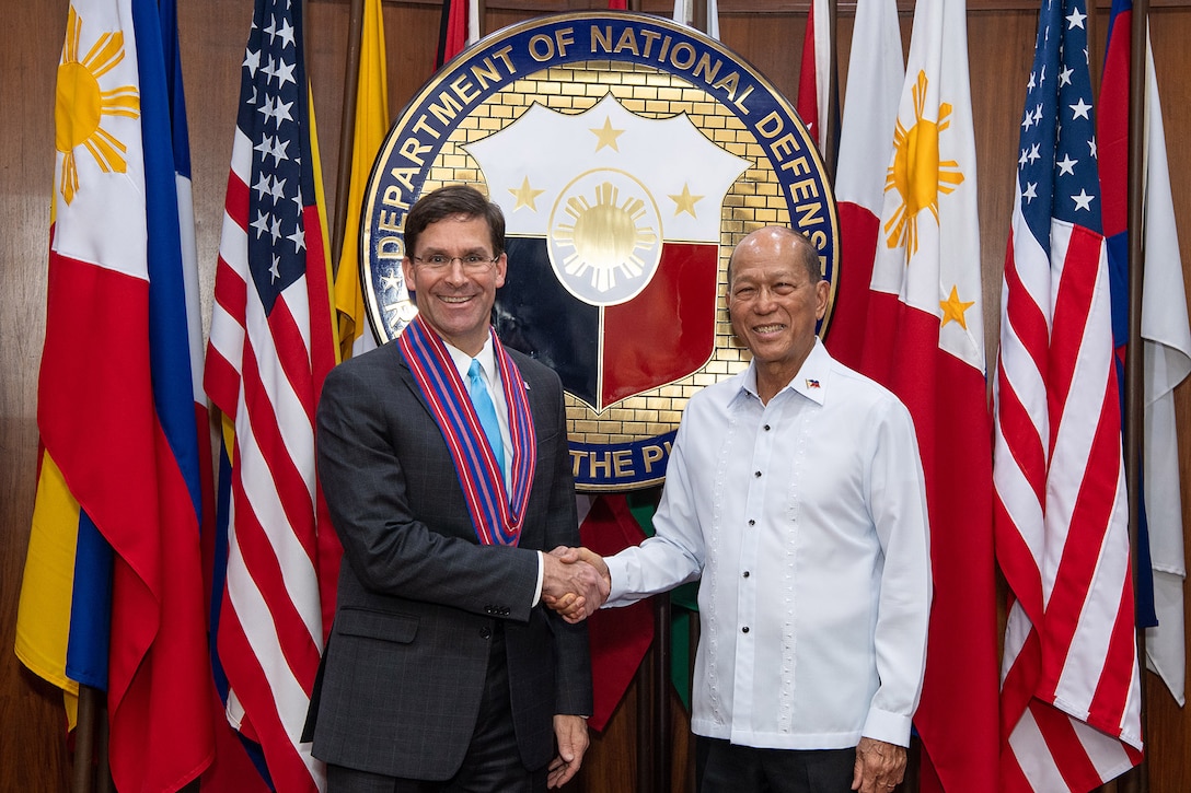 Two men shake hands in front of flags and a Department of National Defense seal.