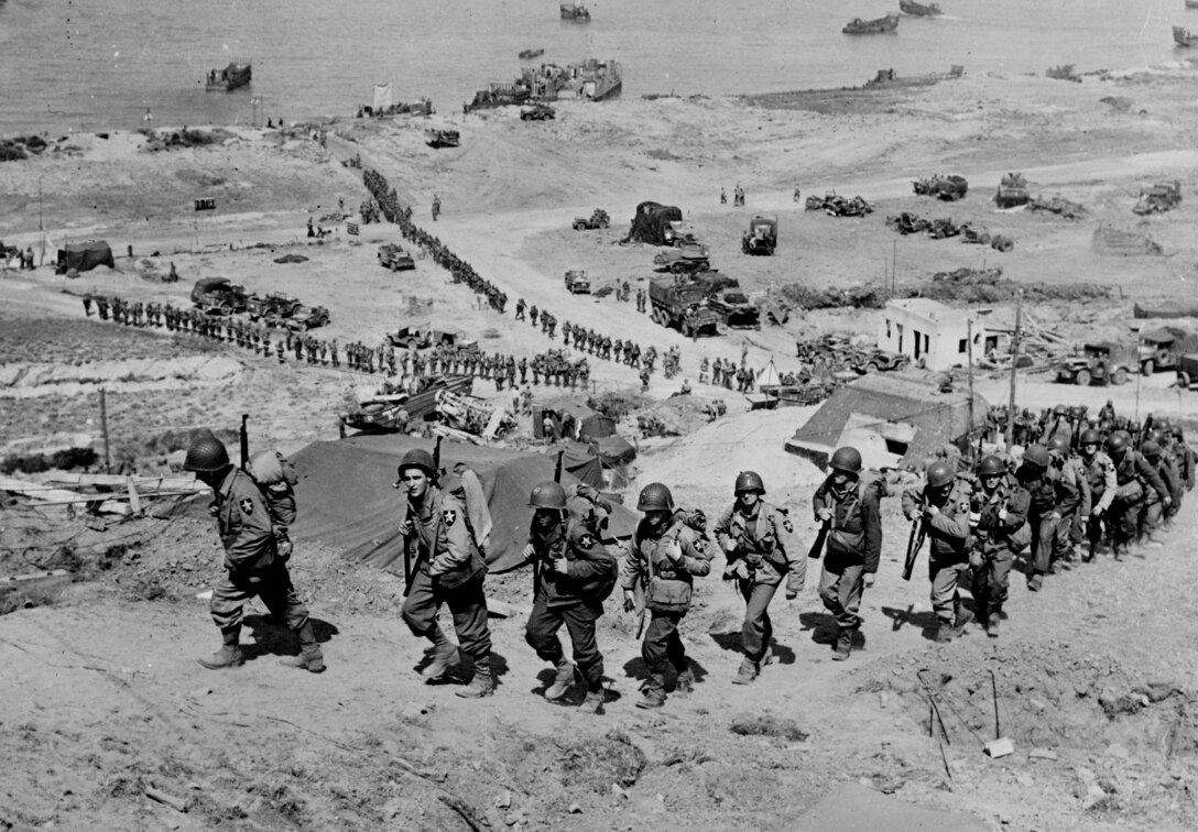 Troops from U.S. Army’s 2nd Infantry Division march up bluff at E-1 draw, passing German bunker Widerstandsnest 65, in Easy Red sector of Omaha Beach, June 7, 1944 (U.S. Army Signal Corps)