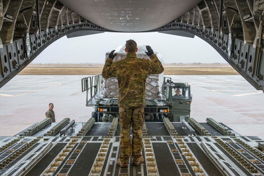 An airman stands inside an open aircraft and guides a vehicle carrying food.