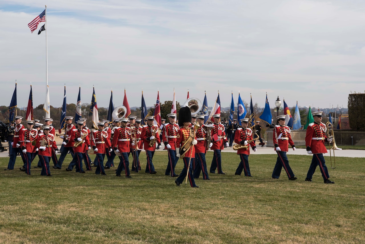 Military band members wearing red jackets and blue trousers and carrying their instruments march in salute.