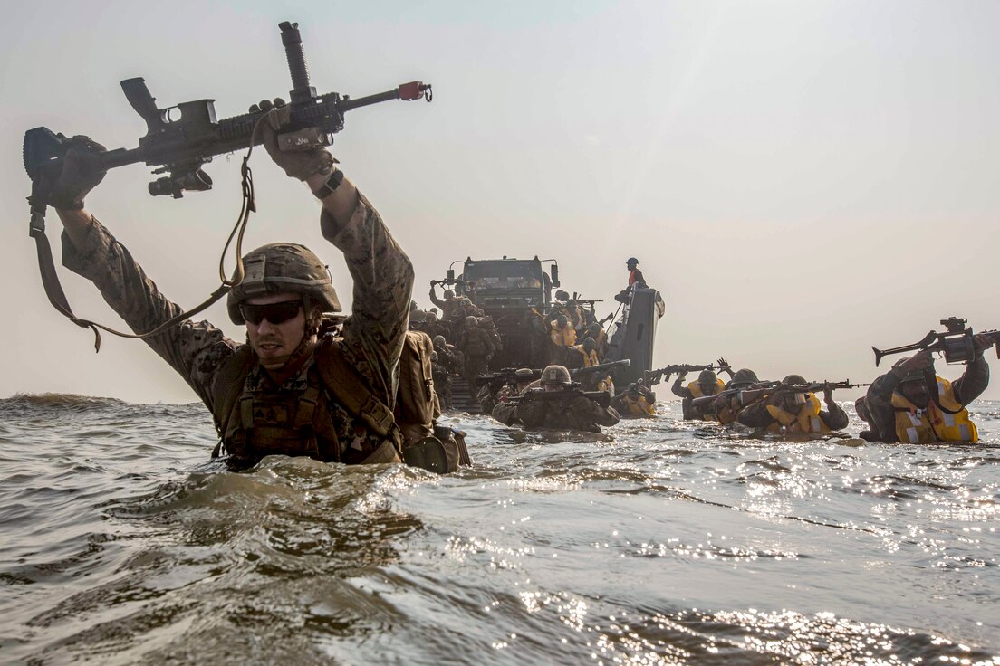U.S. Marines and Indian service members wade in high waters, holding weapons above their heads.