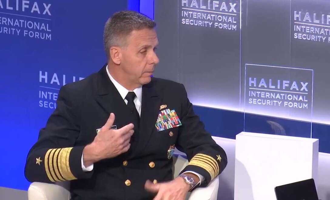 A navy admiral in uniform speaks from a white arm chair.