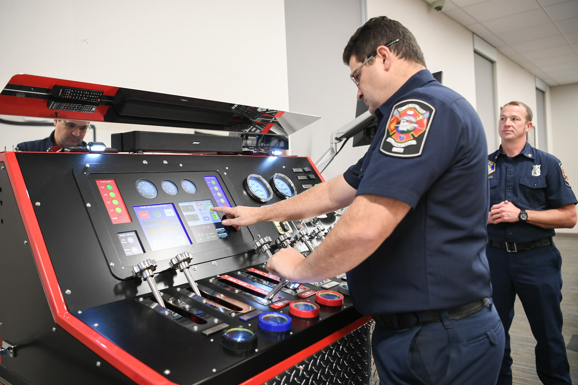 Jason Medina, Fire and Emergency Services (F&ES) driver operator, touches a digital screen on a pump simulator panel that is adorned with handles,dials and gauges, while Daniel Payne, F&ES instructor, observes in the background.
