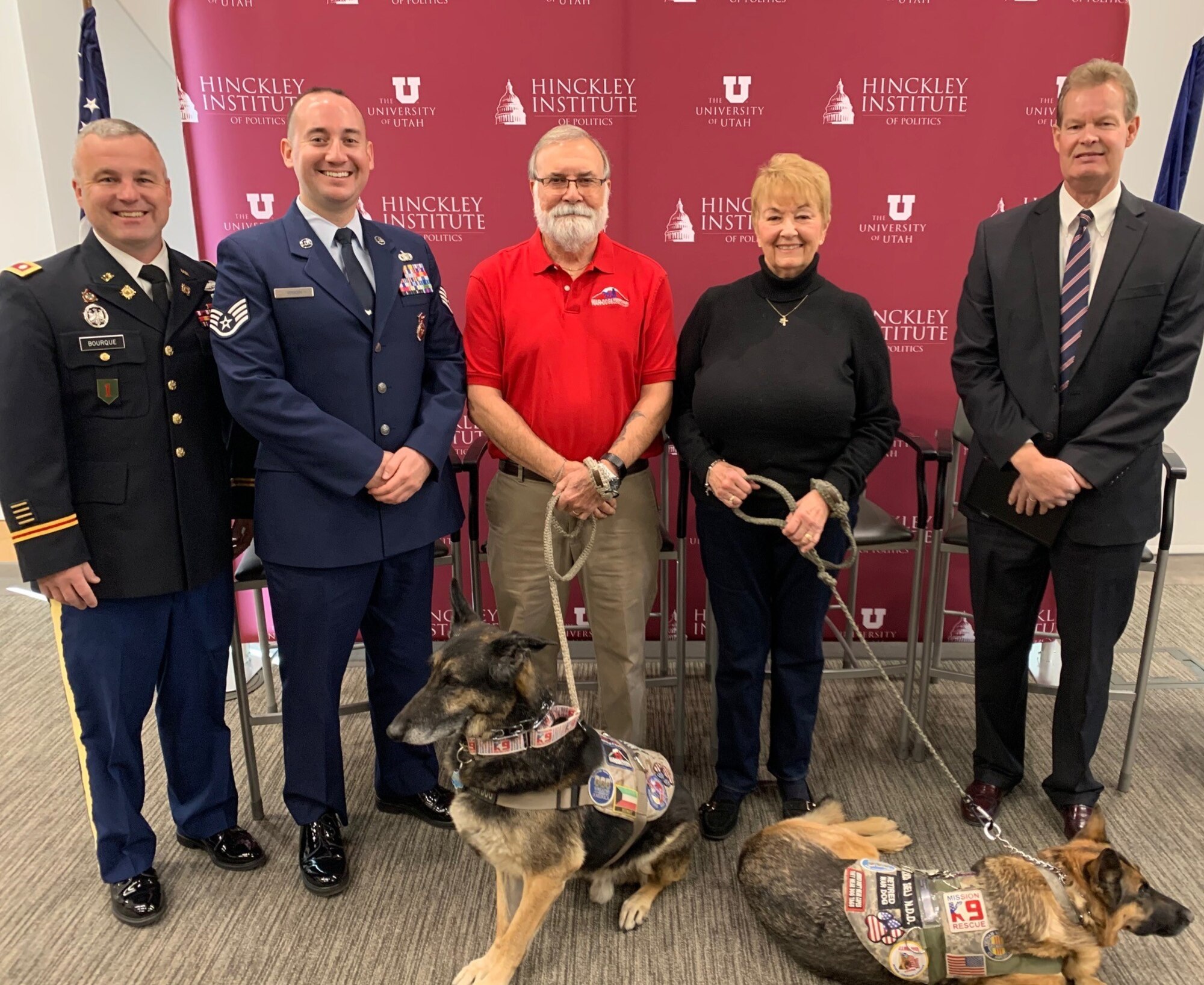 Five panelists and two military working dogs pose in front of a red University of Utah Hinckley Institute for Politics backdrop.