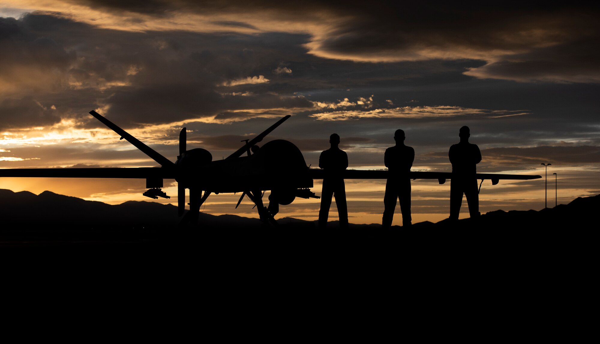 3 Airmen pose in front of an MQ-9 Reaper underneath a Nevada sunset.