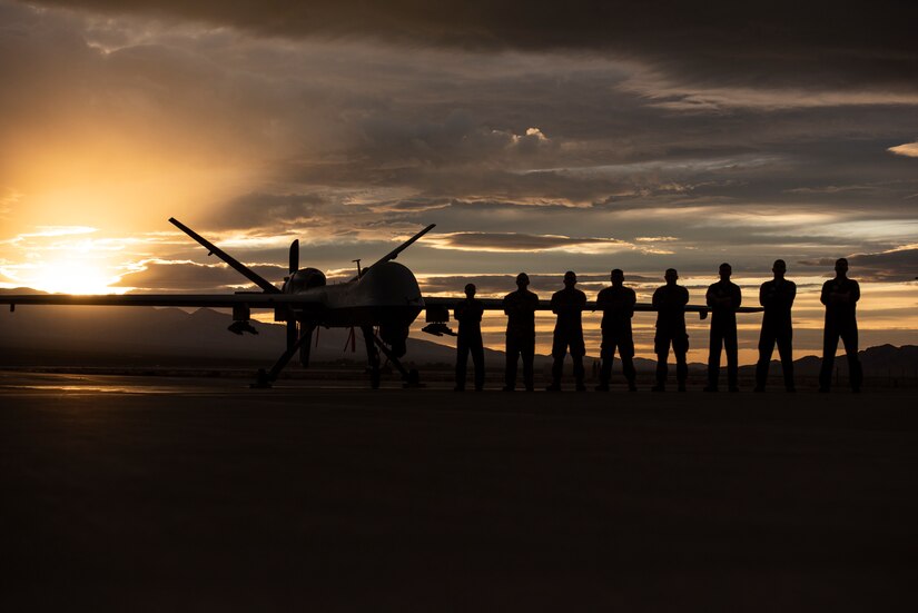 8 Airmen stand in front of an MQ-9 Reaper wing underneath a Nevada sunset.