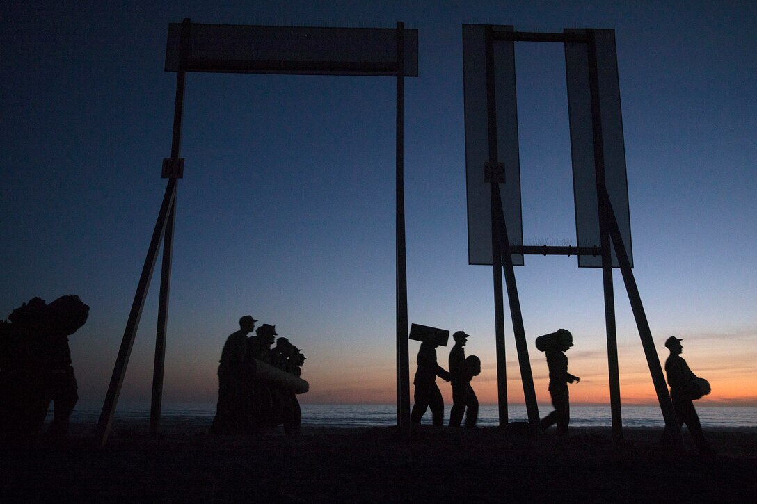 At dusk, service member in silhouette walk along a beach.