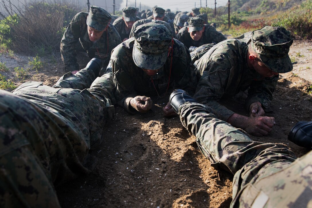 A long line of service members crawl on their bellies through dirt.
