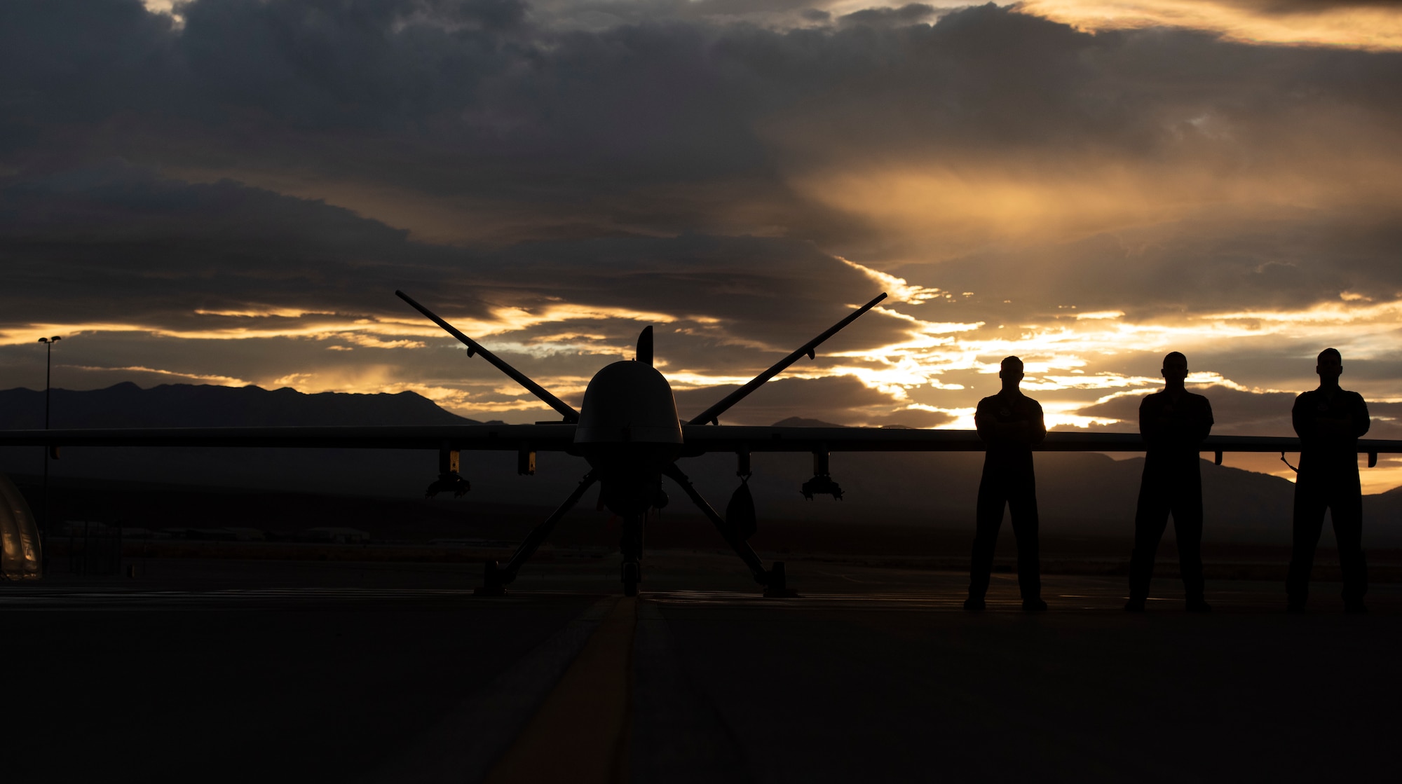 3 Airmen pose in front of an MQ-9 Reaper underneath a Nevada sunset.
