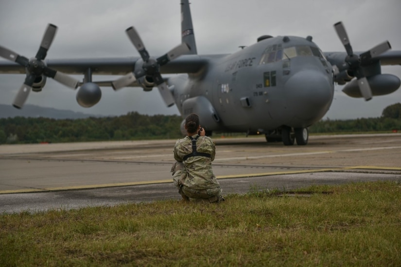 A soldier kneels on the tarmac to take photos of a C-130H Hercules aircraft.
