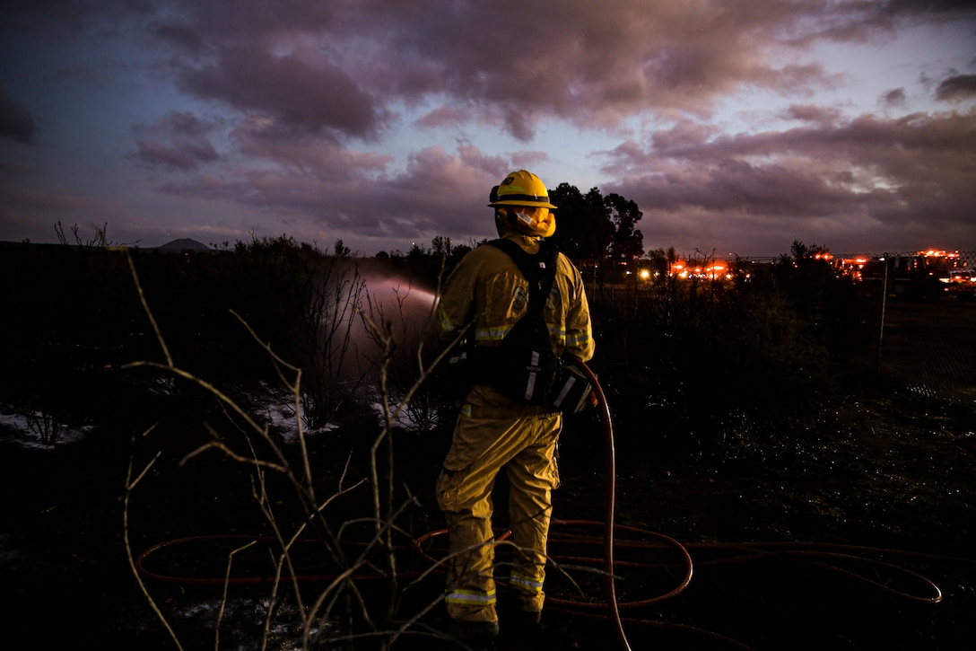 A Marine Corps firefighter uses a hose to put out a fire at twilight.