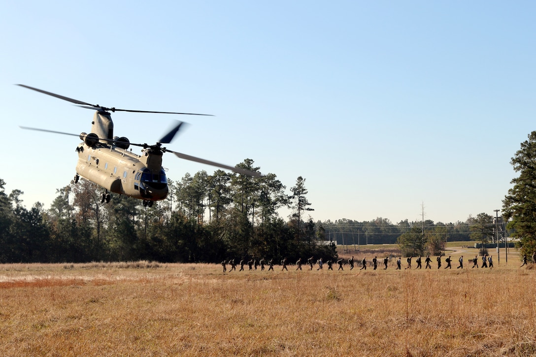 A line of soldiers walk away as a helicopter lifts off from a field.