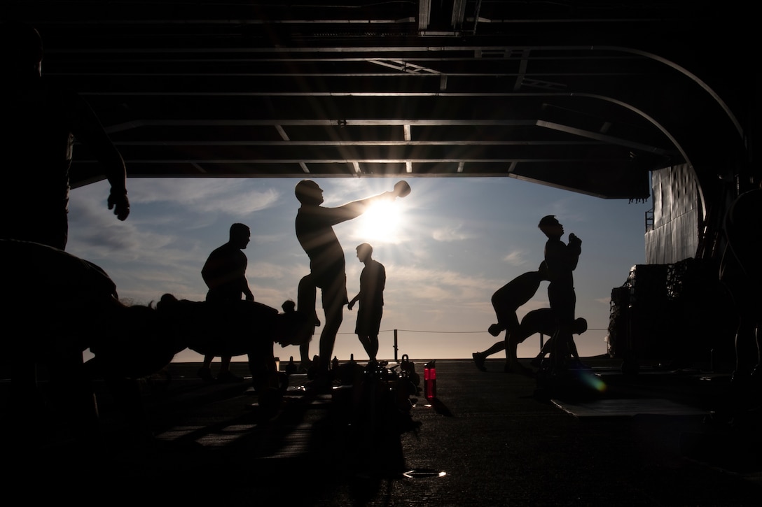 Silhouettes of sailors doing exercises in the hangar bay of a ship.