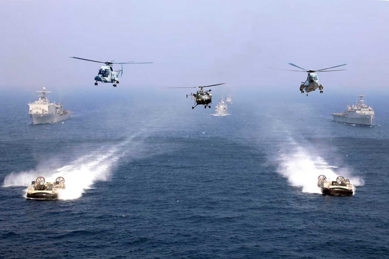 Three helicopters fly above two air-cushion landing crafts and three ships.