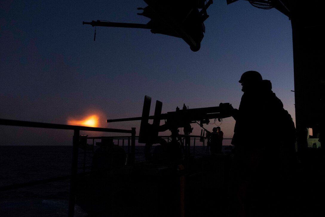 Sailors fire a machine gun from a military ship at twilight.