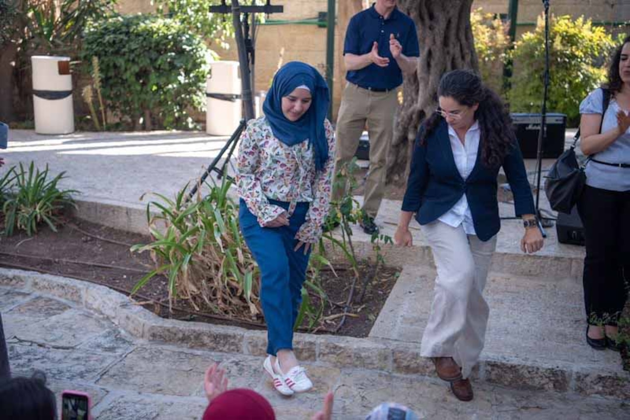 Two women, one teaching and one learning, lift a foot up as they look down at their feet while dancing. A person in the background is clapping.