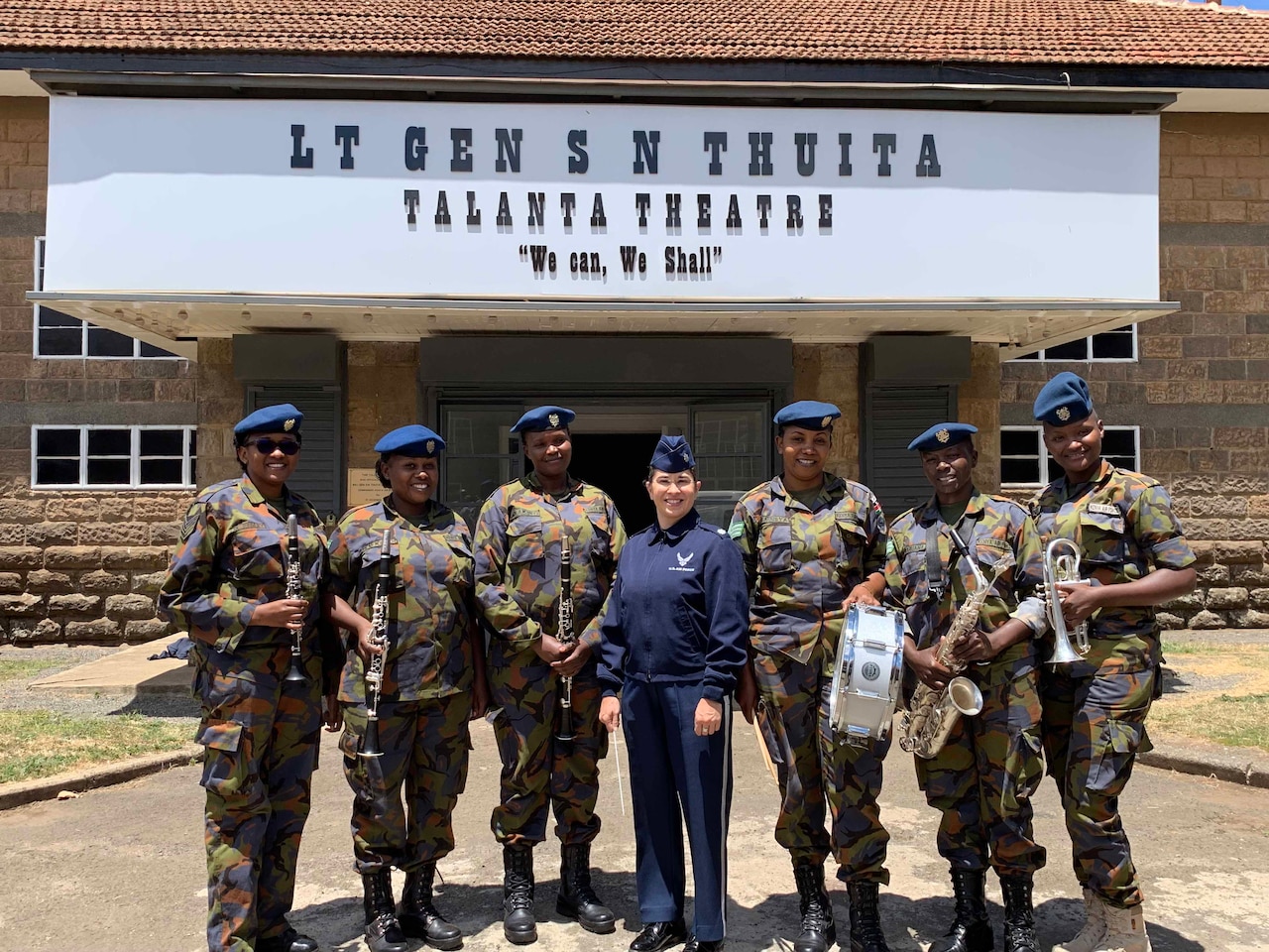 A female U.S. Air Force member poses with six Kenyan Air Force Band members, all of whom are holding instruments, outside a theater.