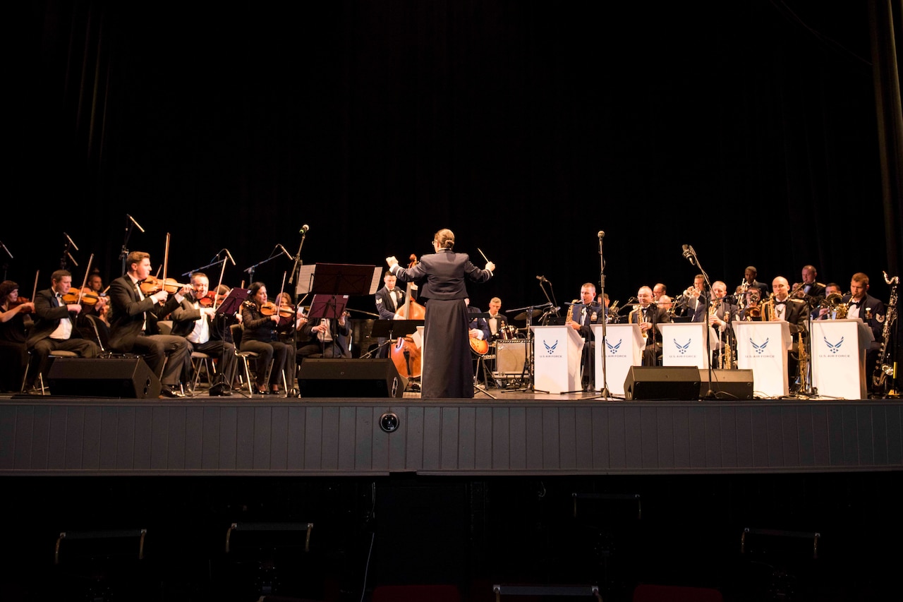 A conductor leads a large band with woodwind and brass instruments plays on stage.