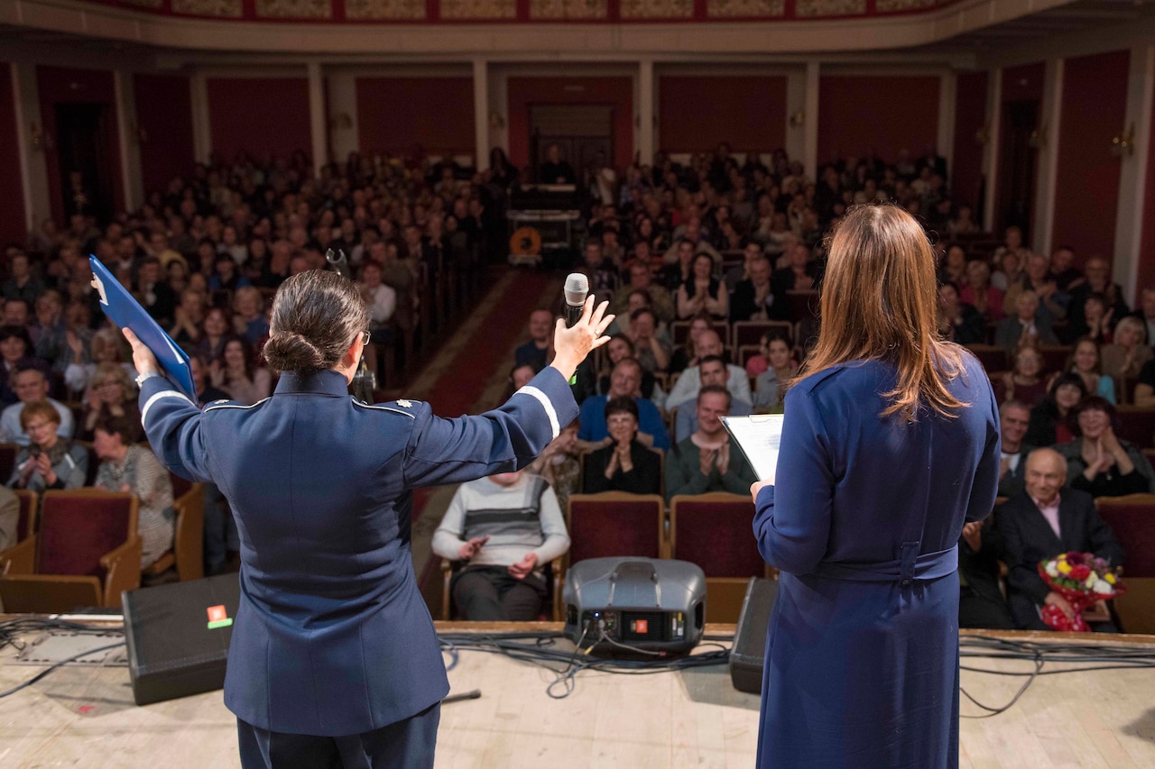 Two people on a stage stand facing a crowd, one with her arms open wide, to greet a theater audience.