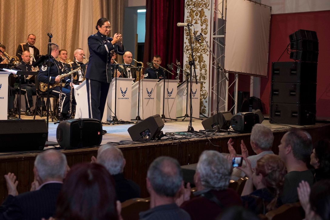 A female conductor on stage clasps her hands as theatergoers clap for her. Musicians sit in chairs behind the conductor.