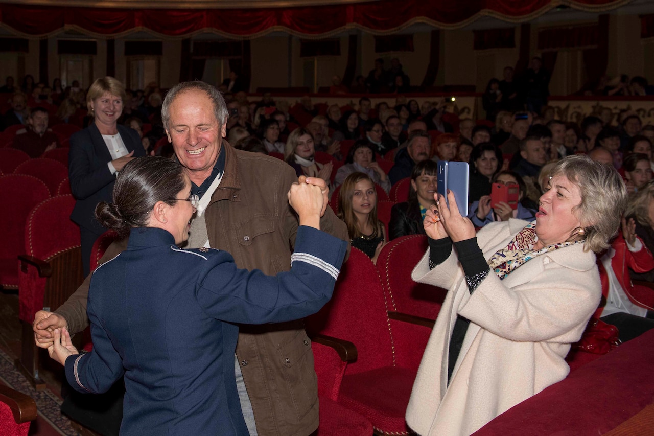 An Air Force band conductor dances with an audience member in the aisle of a theater as another theatergoer uses her phone to snap a photo.