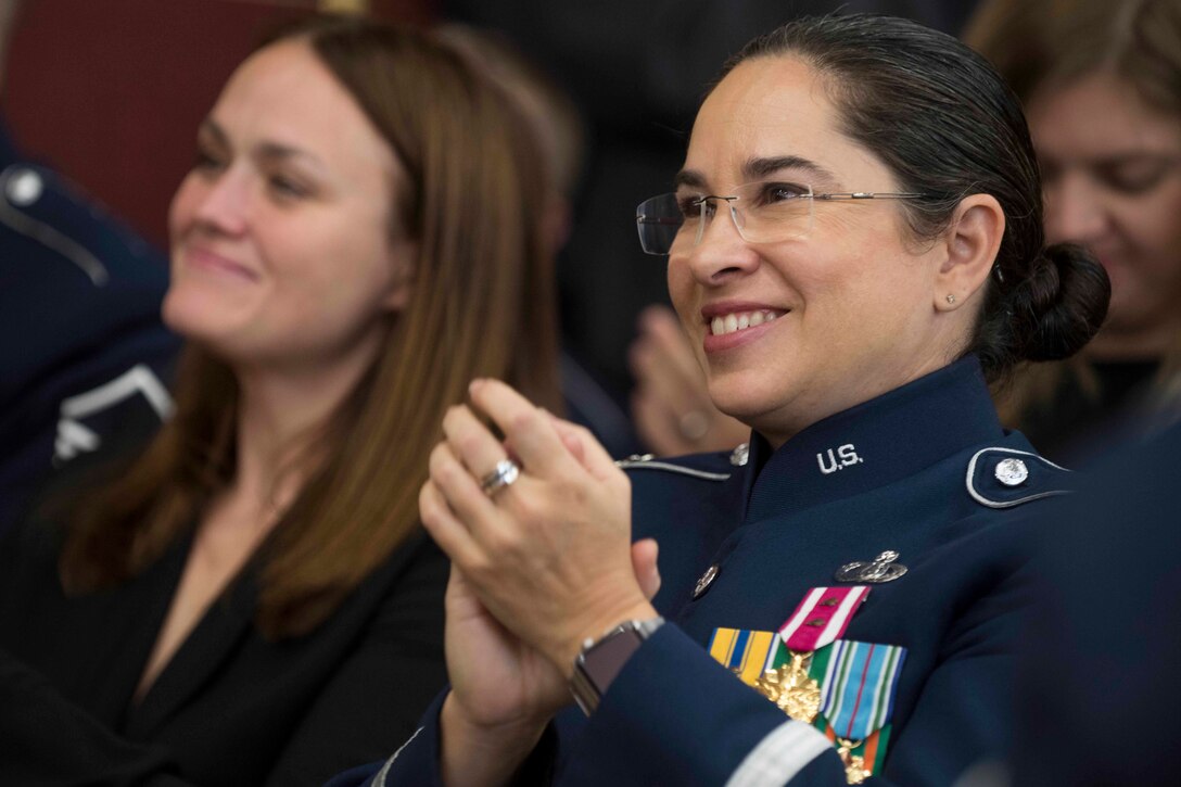 A female Air Force lieutenant colonel in dress blue uniform applauds in a crowd as someone smiles beside her.