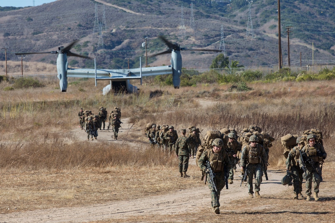 A group of Marines walk in a field with a helicopter in the background.