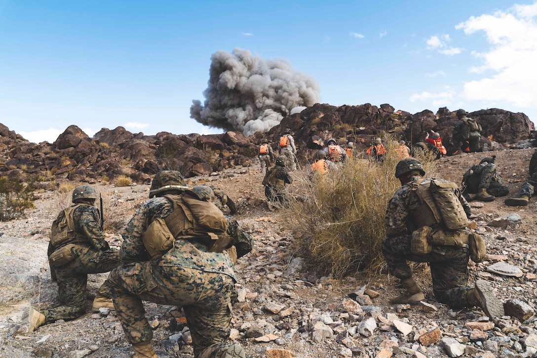 Marines kneel in a desert terrain as smoke rises in the distance.