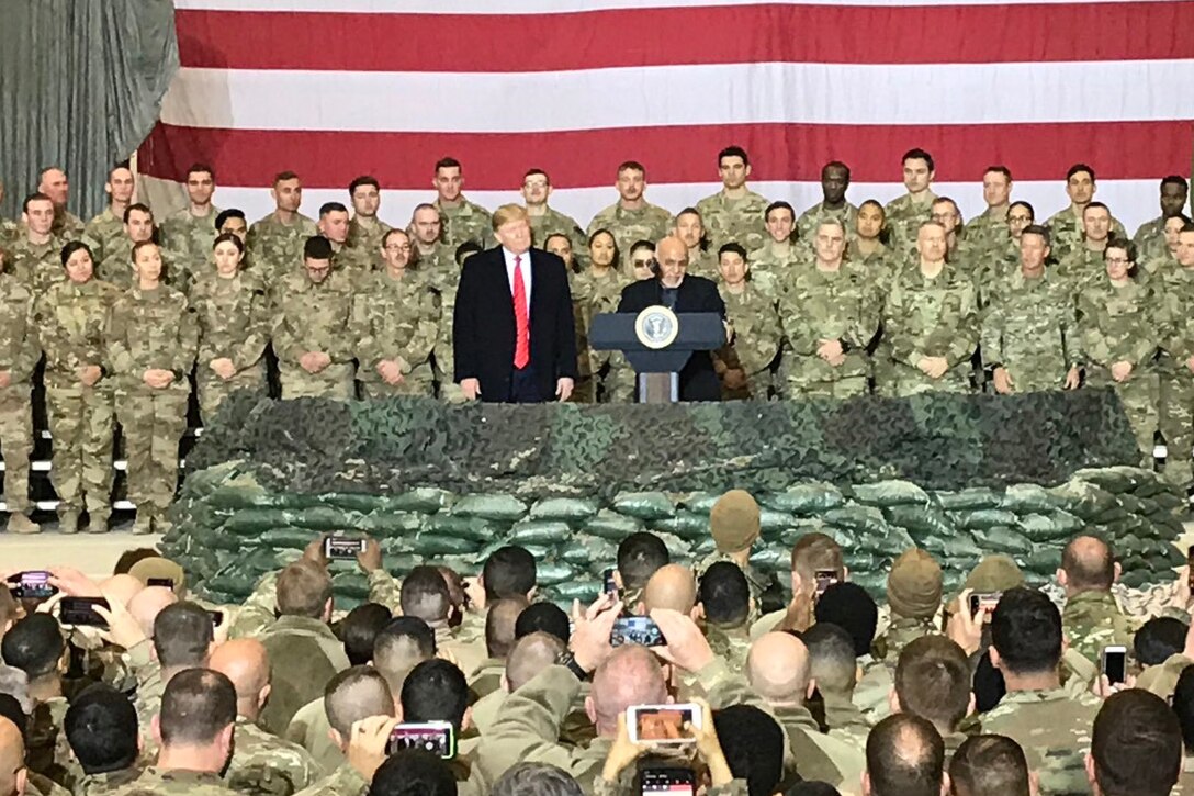 Two men stand on a stage surrounded by members of the military.