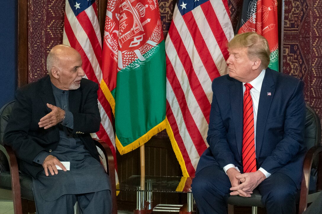 Two leaders sit in chairs and talk with flags behind them.