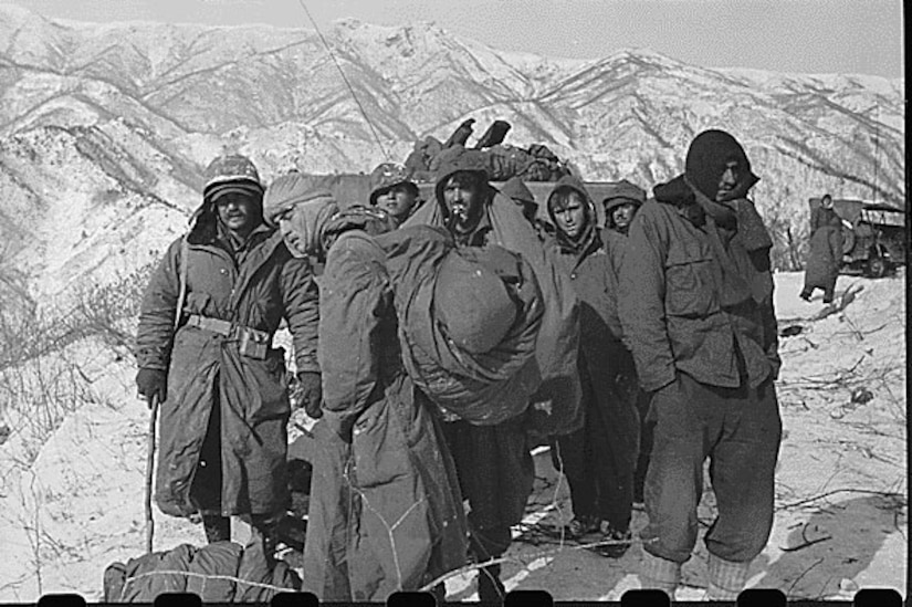 Several men in parkas and heavy clothing stand around together, with snow-covered mountains in the background.