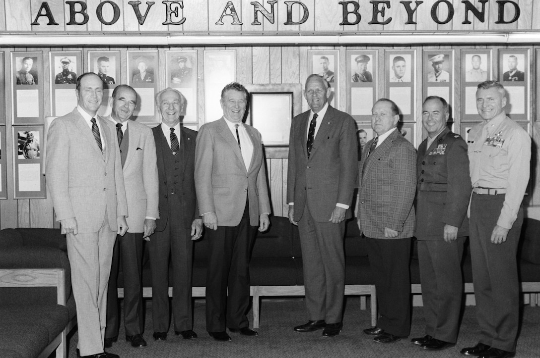 Eight Medal of Honor recipients pose in front of a wall of plaques and photos with the words “Above and Beyond.”