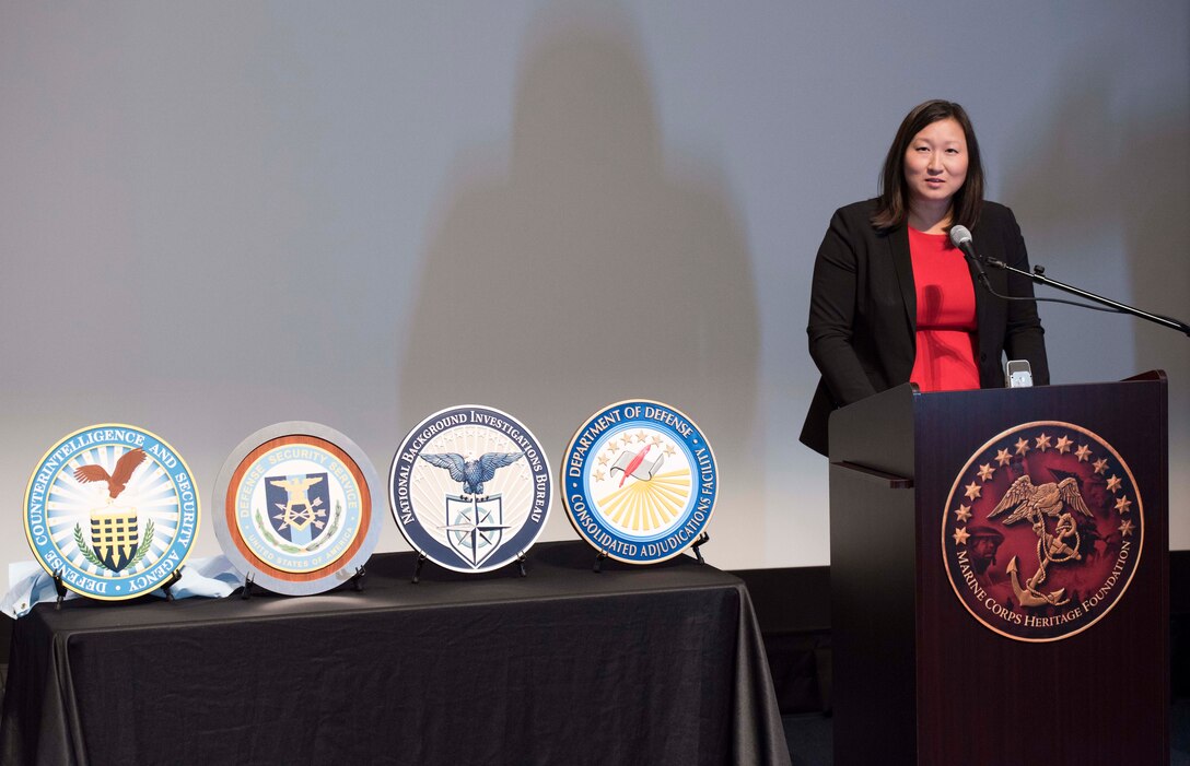 Woman stands behind a lecturn; to her right is a table with the emblems of four agencies.