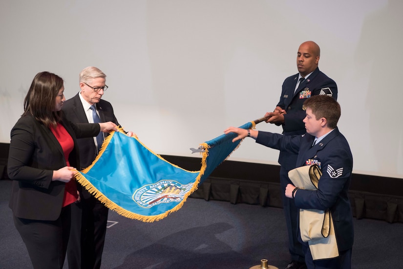 Four people, including two in uniform, unfurl a turquoise flag trimmed in gold fringe.