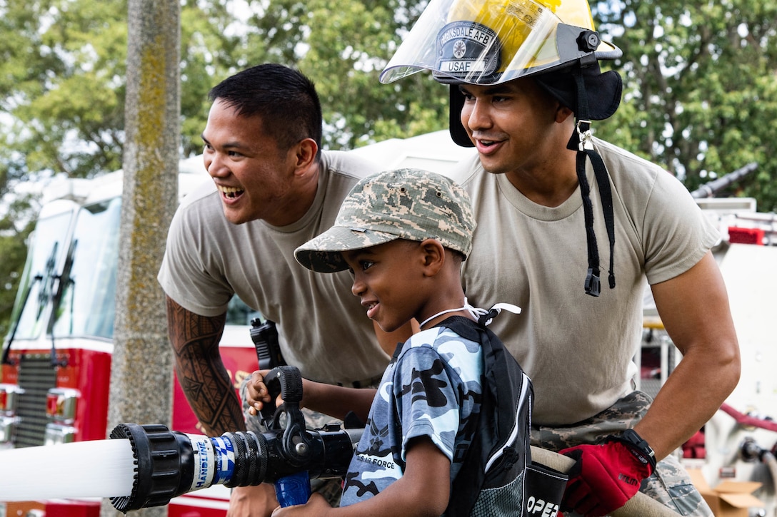 Two airmen smile as they help a boy operate a water hose.